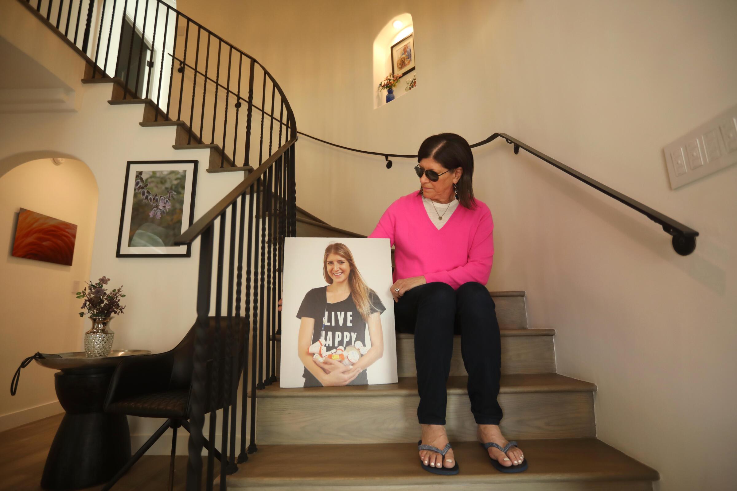 A woman in sunglasses and a pink top sits on a staircase holding a portrait of a young woman whose shirt reads "Live Happy." 