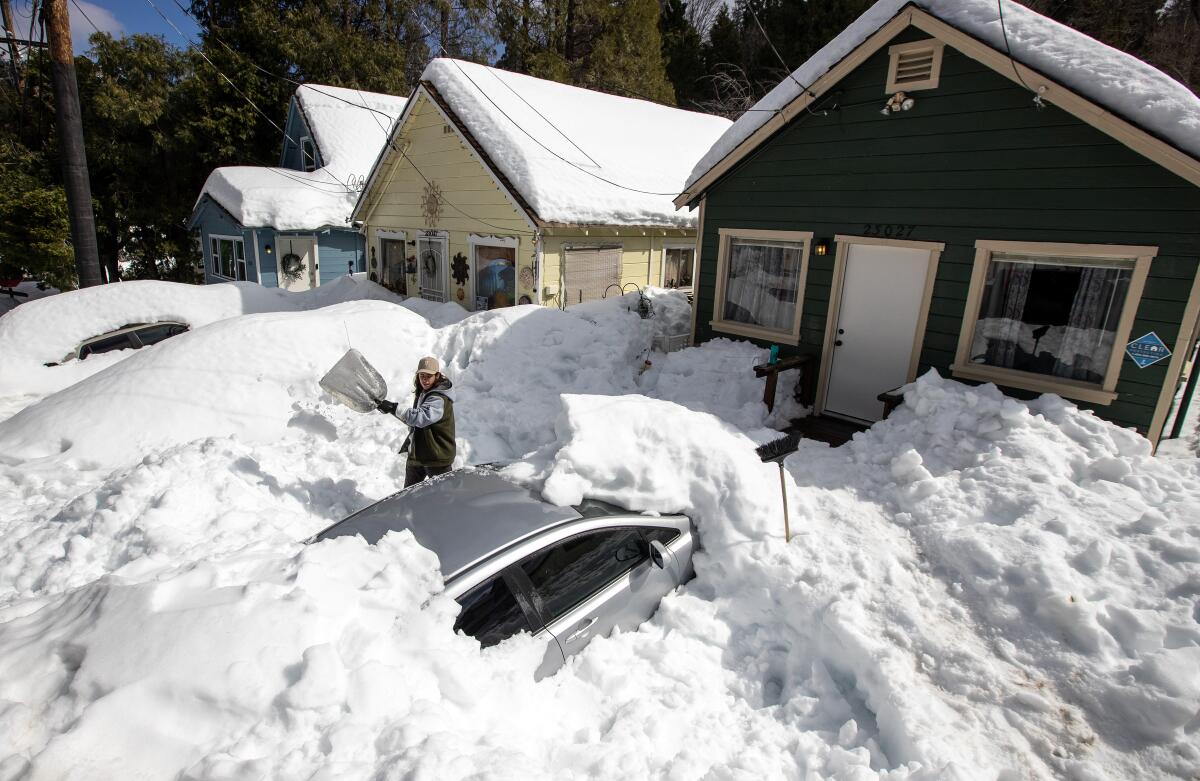 A person shovels snow that is covering a car and several houses.
