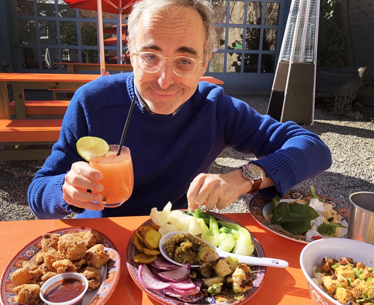 Author Gary Shteyngart sitting at an outdoor table with several plates of food, holding a drink.