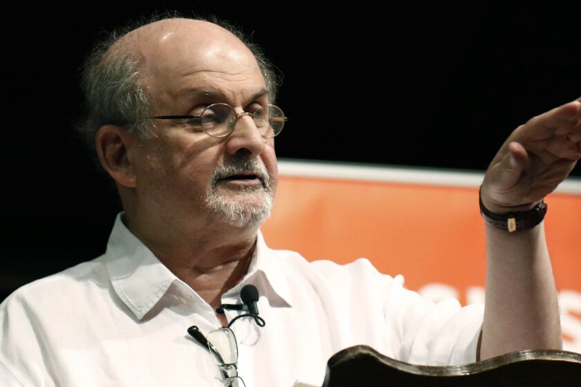 A balding man with a gray goatee and glasses gestures with his left hand while speaking to an unseen audience