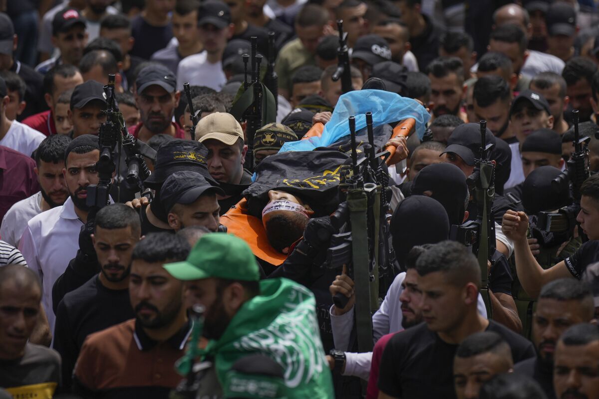 A body is carried on a stretcher above a crowd of mourners, some of whom are armed.