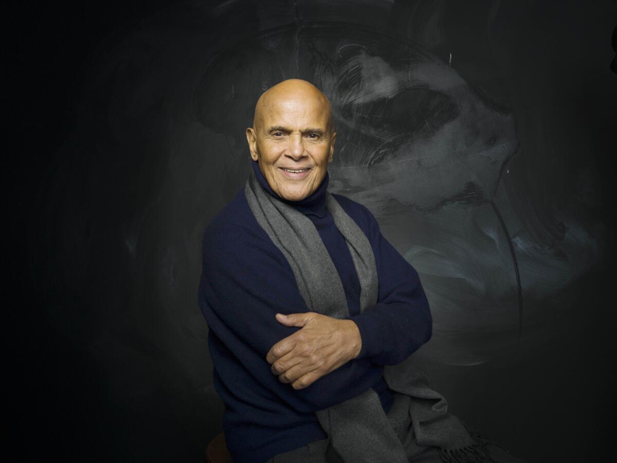 Harry Belafonte wearing a dark sweater with a gray scarf around his neck and his arms folded