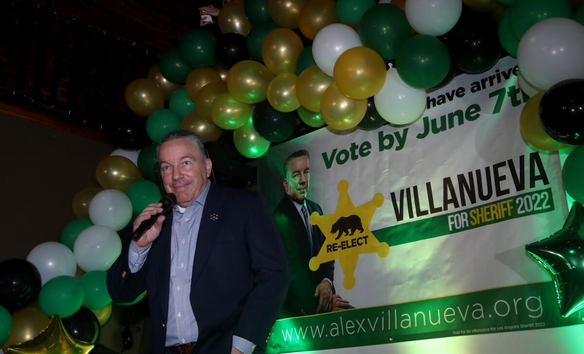 A man speaks in front of a balloon arch and a "Villanueva" banner.
