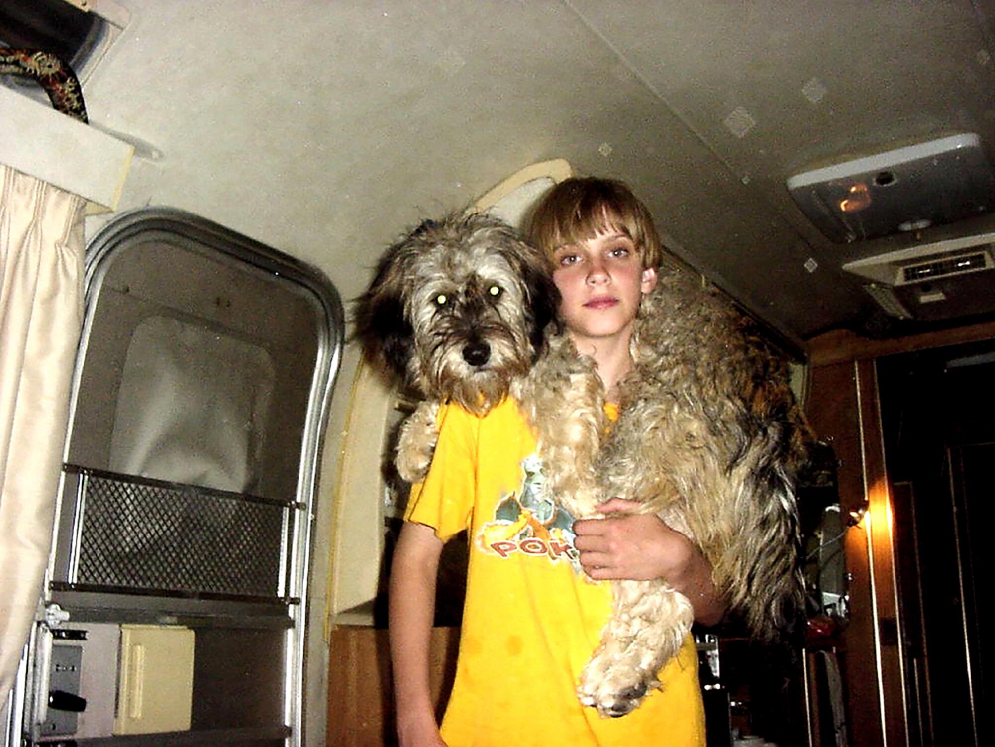 A childhood photo of Zo? Bossiere, in a yellow T-shirt, standing in an RV with a dog on their shoulders.
