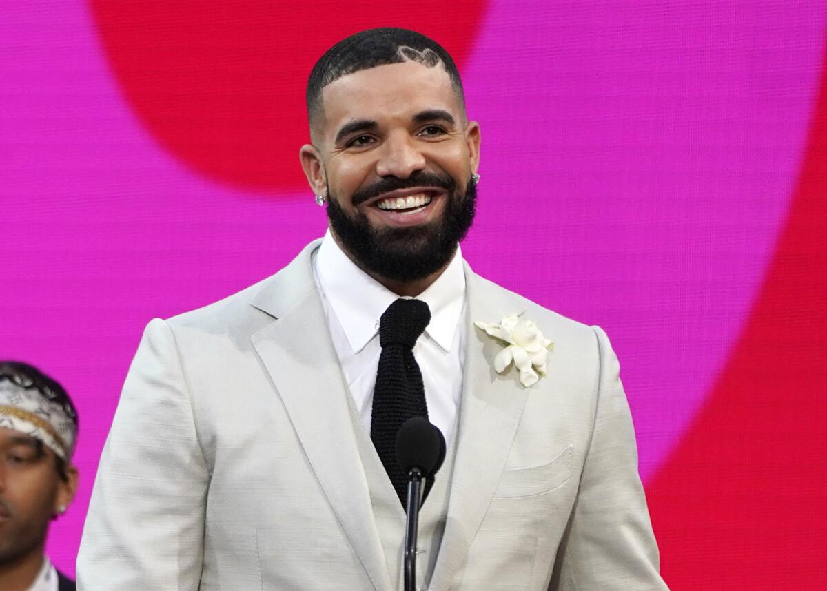 Drake smiling while wearing a light gray suit and black tie in front of a red and hot pink backdrop