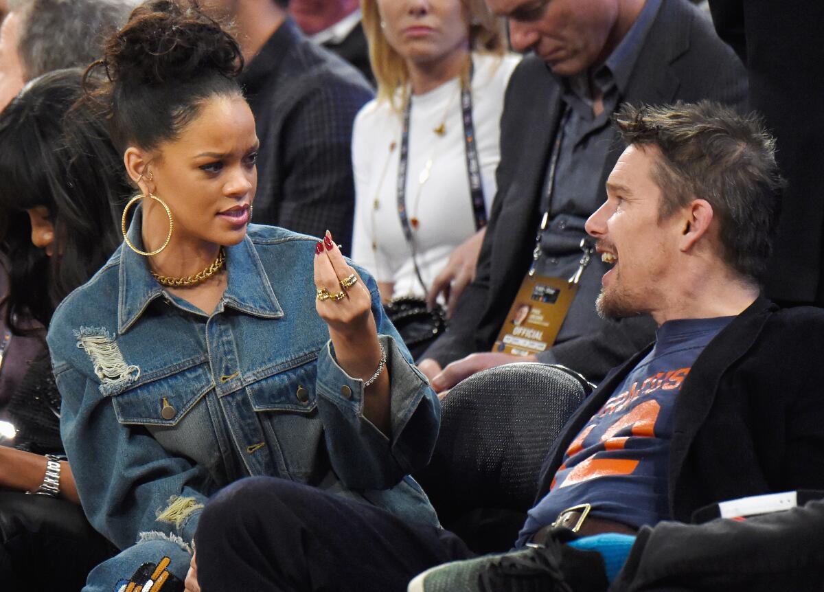 Rihanna in a jean outfit speaking to Ethan Hawke in a dark blazer and graphic t-shirt at a basketball game