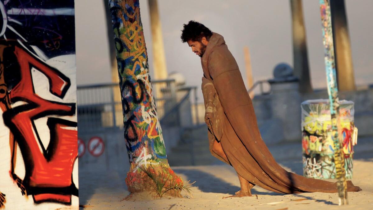 At dawn, a homeless man walks around Venice Beach wearing a blanket.