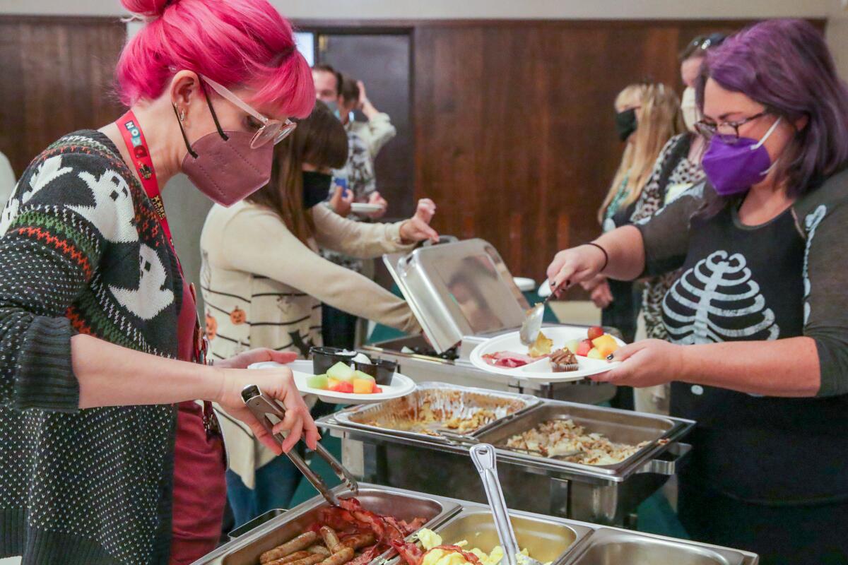 A woman with pink hair grabbing sausage with tongs to put on her plate in a buffet line.