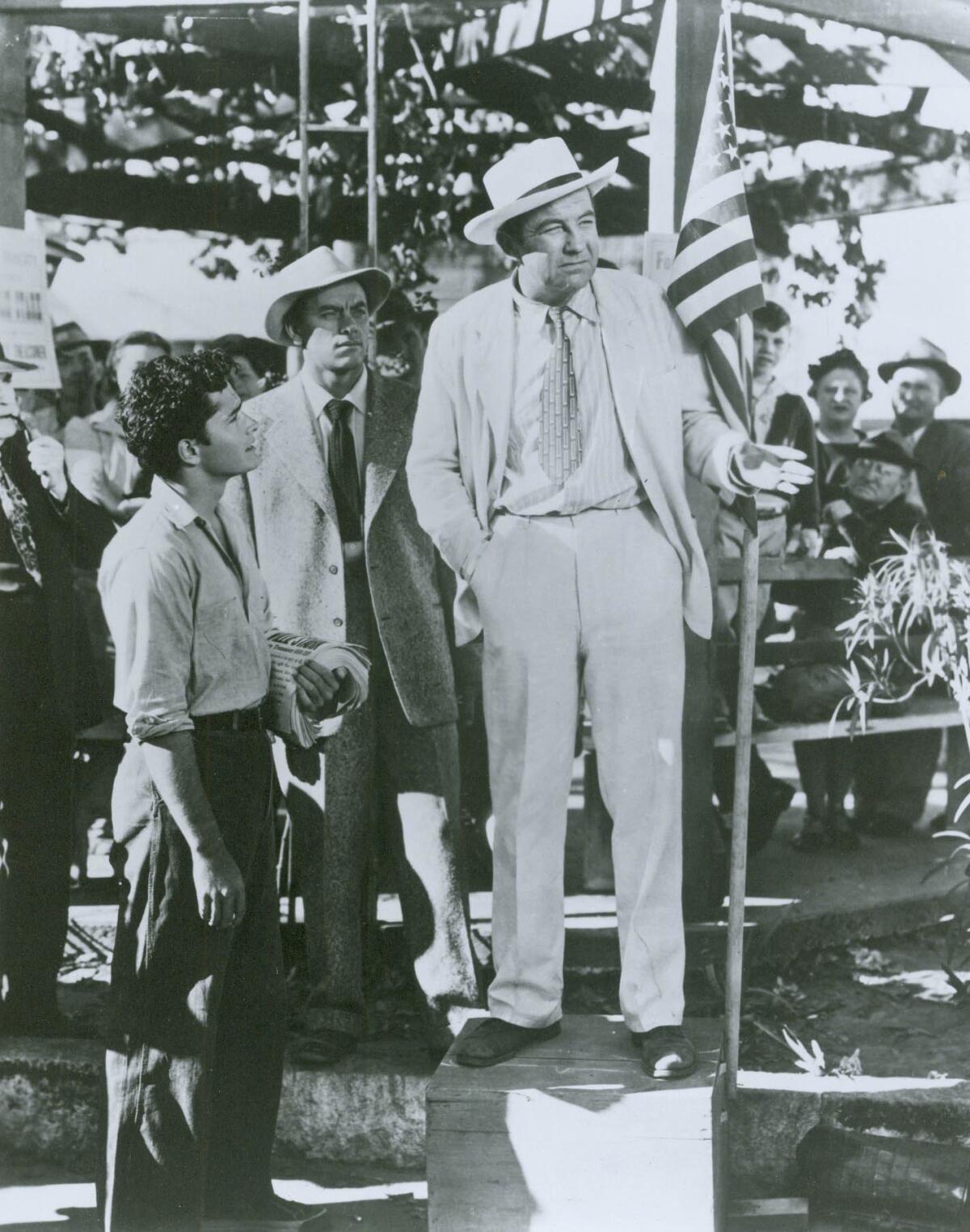 A black-and-white movie still of two men looking at another man in a light suit, standing outside, with a crowd behind them.