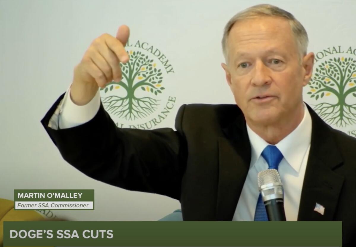 A gray-haired man in dark suit and blue tie points down with his hand while speaking before a microphone