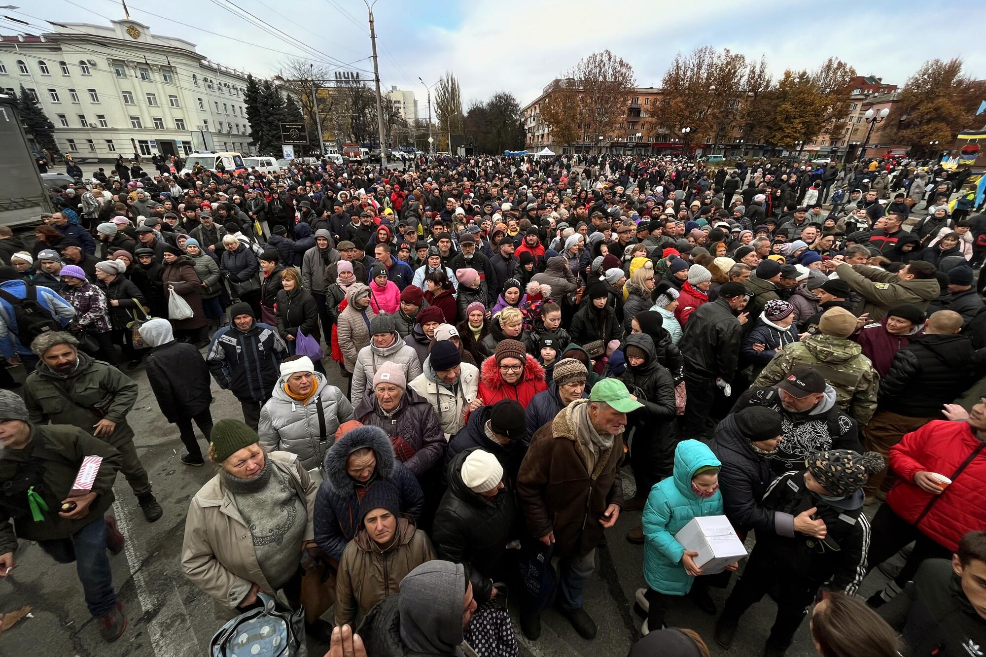 A large crowd of people wearing heavy winter clothing and hats stand near buildings