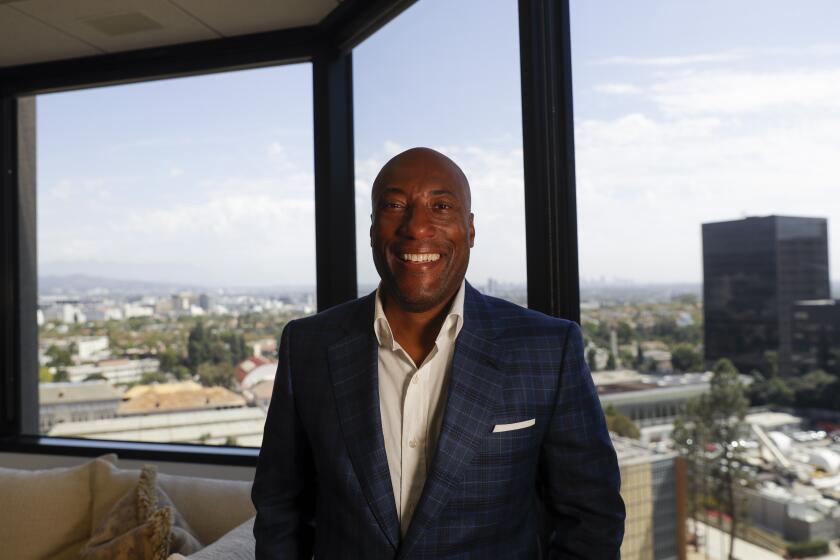 Byron Allen smiling in a dark blue suit in an office with a window view overlooking Los Angeles