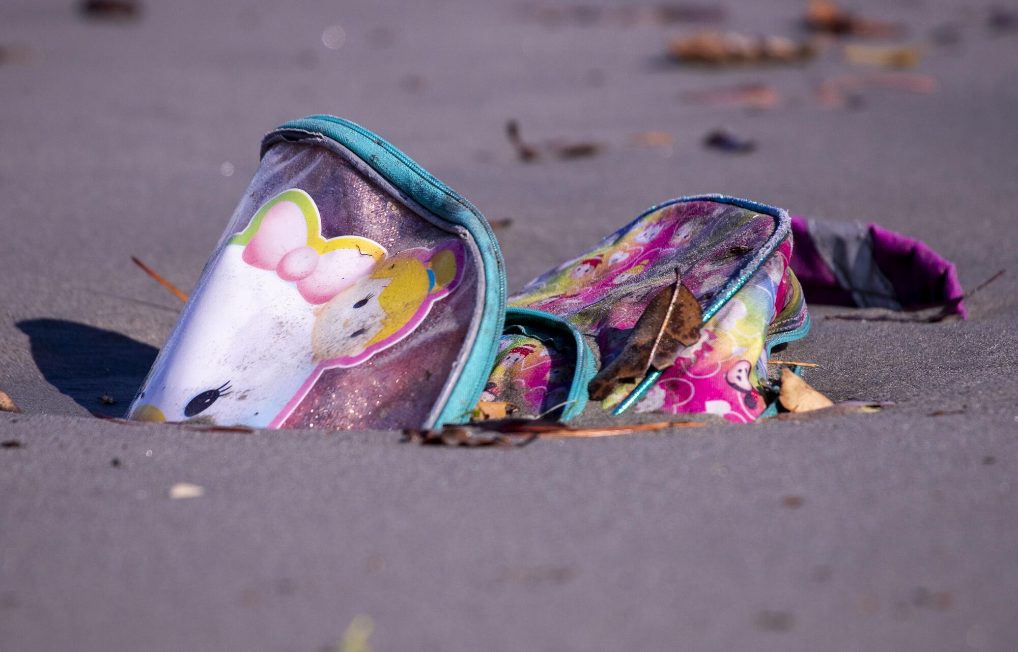 A child's backpack is partially buried in the sand amid a wide swath of trash