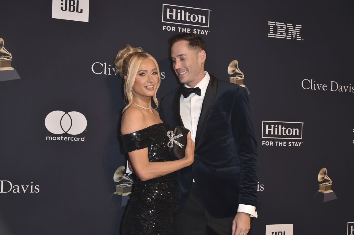 A blond woman with an updo and a sparkly black dress stands arm in arm with a man wearing a black tuxedo