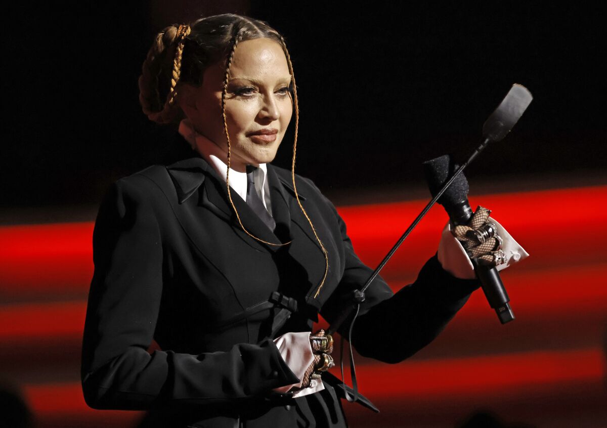 A woman with skinny braids and a black suit holds a microphone in one hand and a riding crop in the other at an awards show