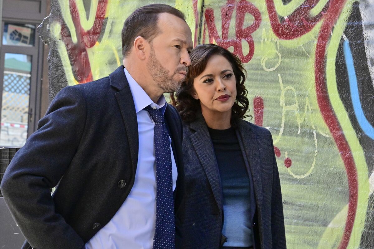 A man and a woman in dark suits stand before a decoratively painted and graffitied wall.