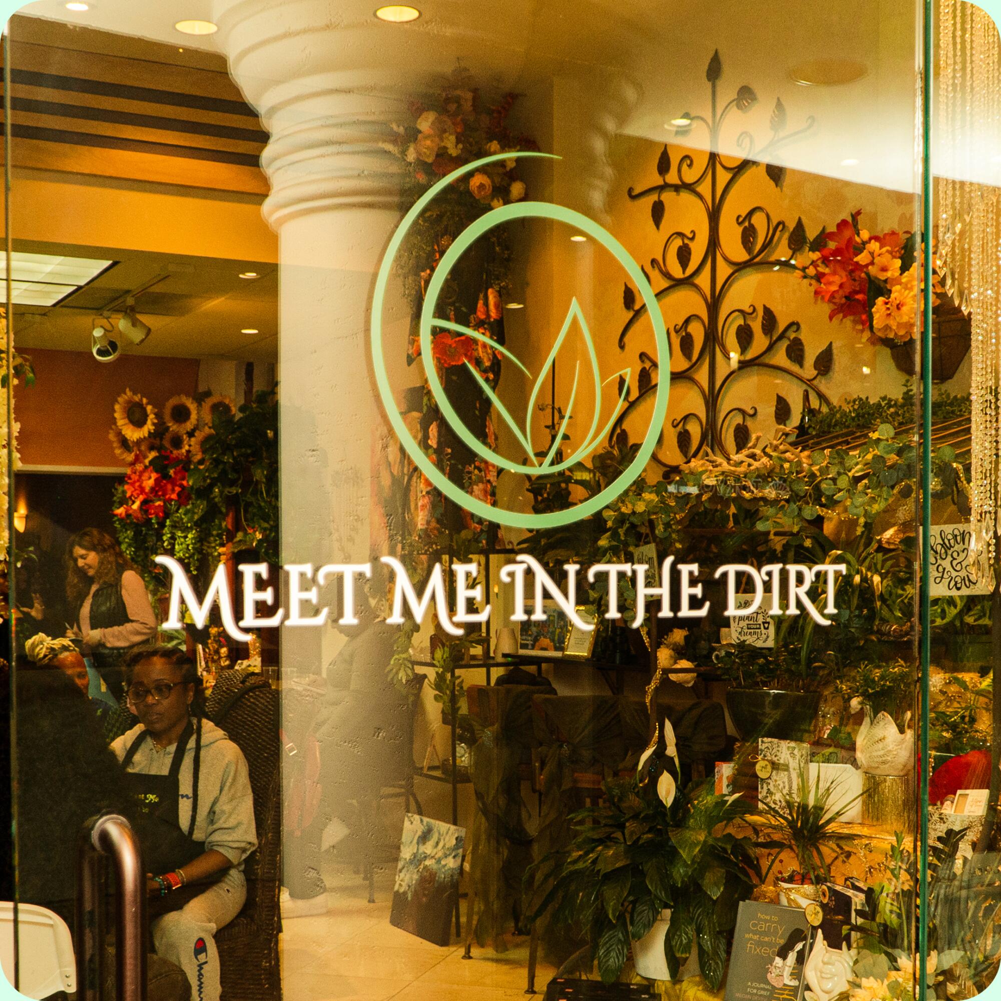 The logo and text for "Meet Me in the Dirt" is seen on a window. People and flowers are seen inside.