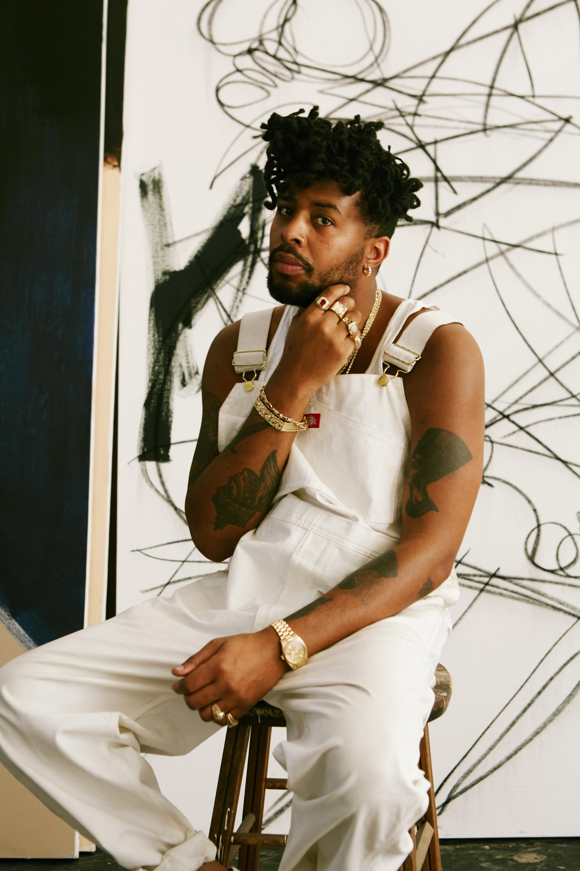 An artist wearing white coveralls sits on a stool in front of an artwork