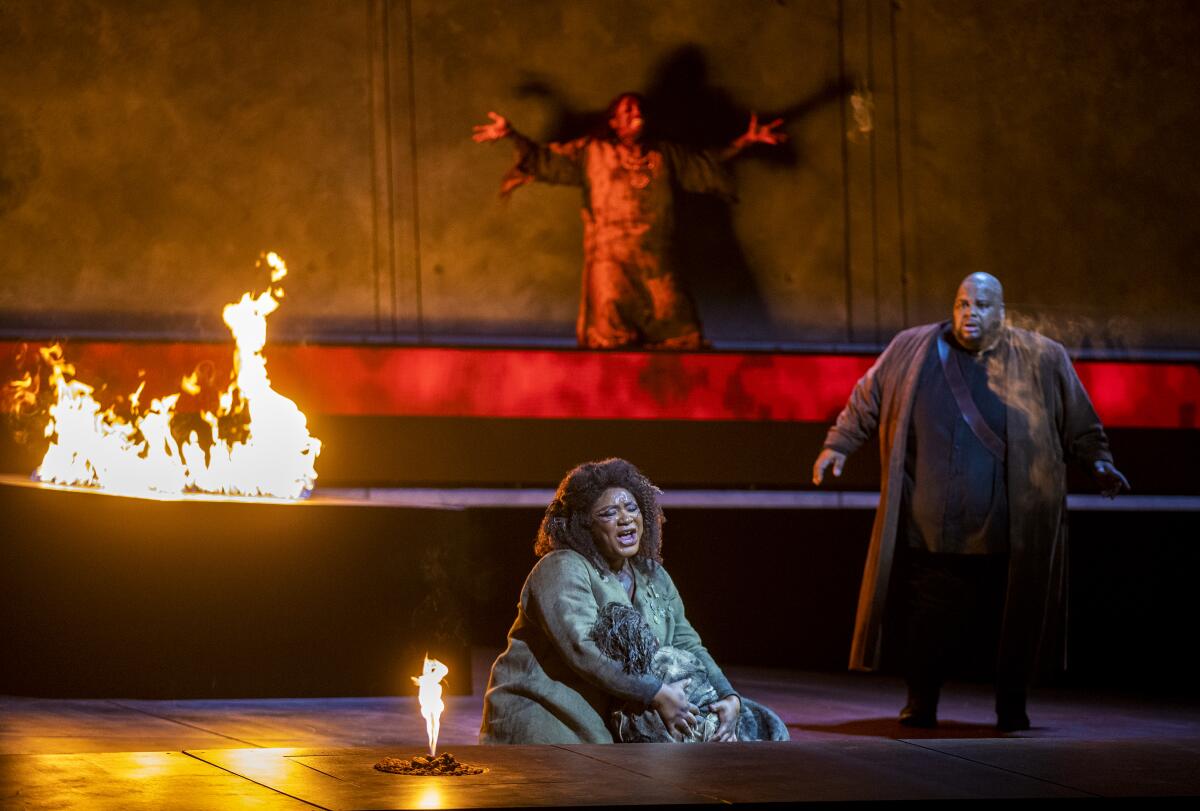 A woman sits on the stage, singing and holding a child while fire burns on the set
