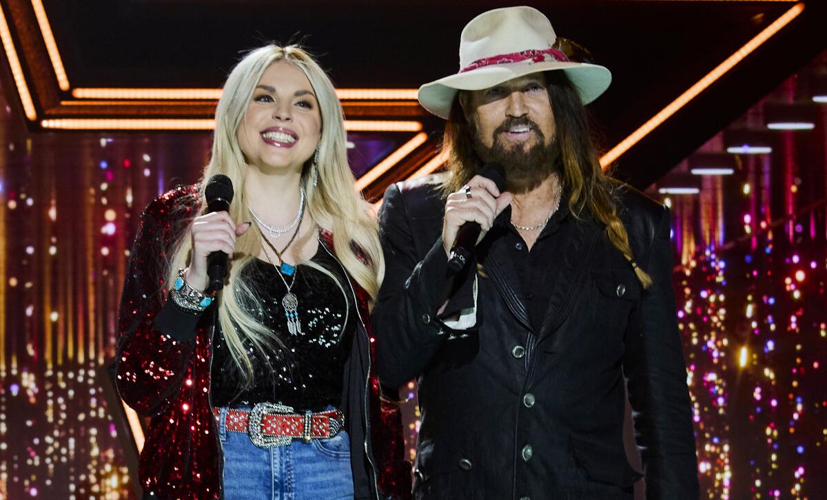 Firerose smiling and holding a microphone next to a bearded Billy Ray Cyrus, who is wearing a hat and speaking into a mic