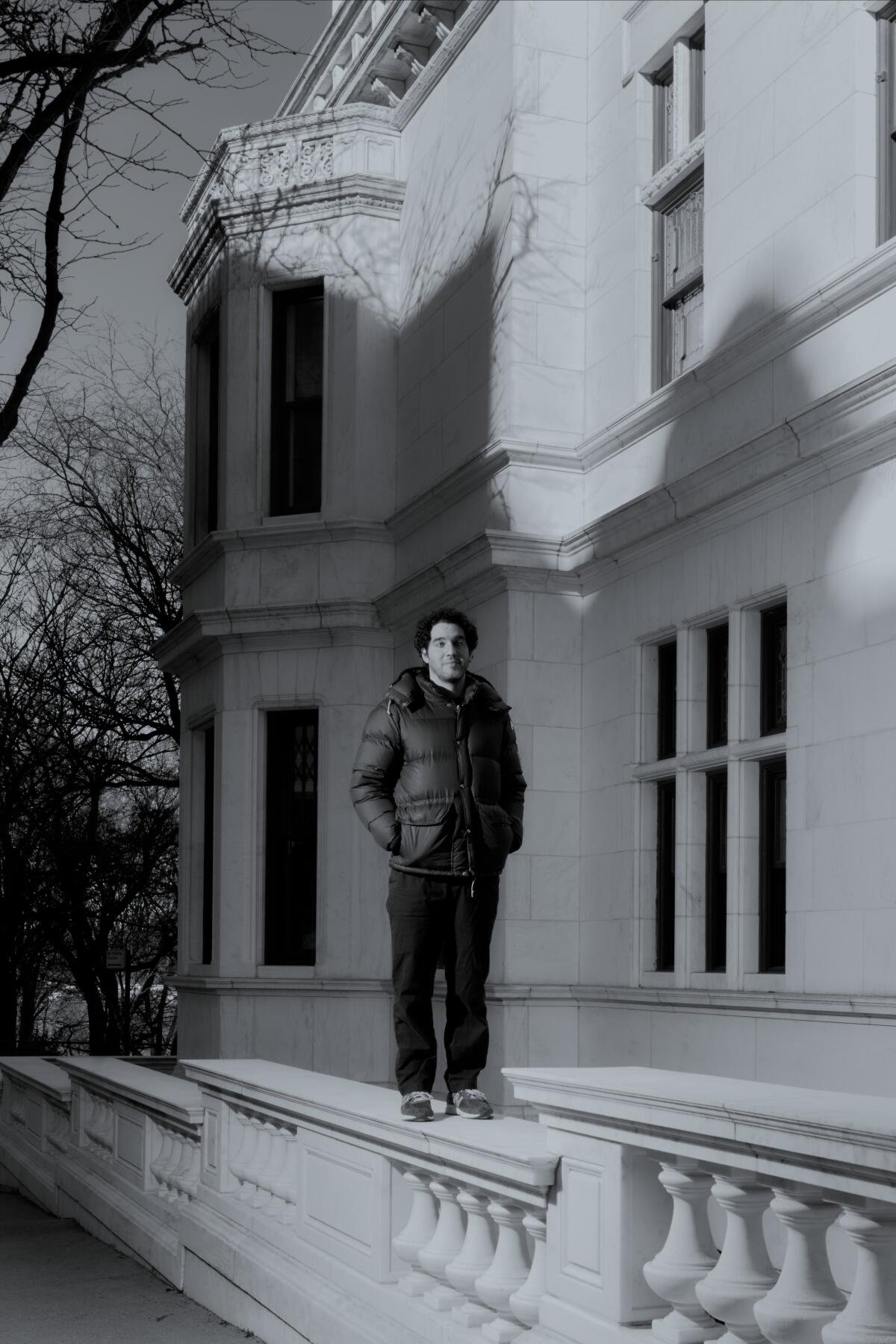Benny Safdie stands on a wide carved railing outside a townhouse.