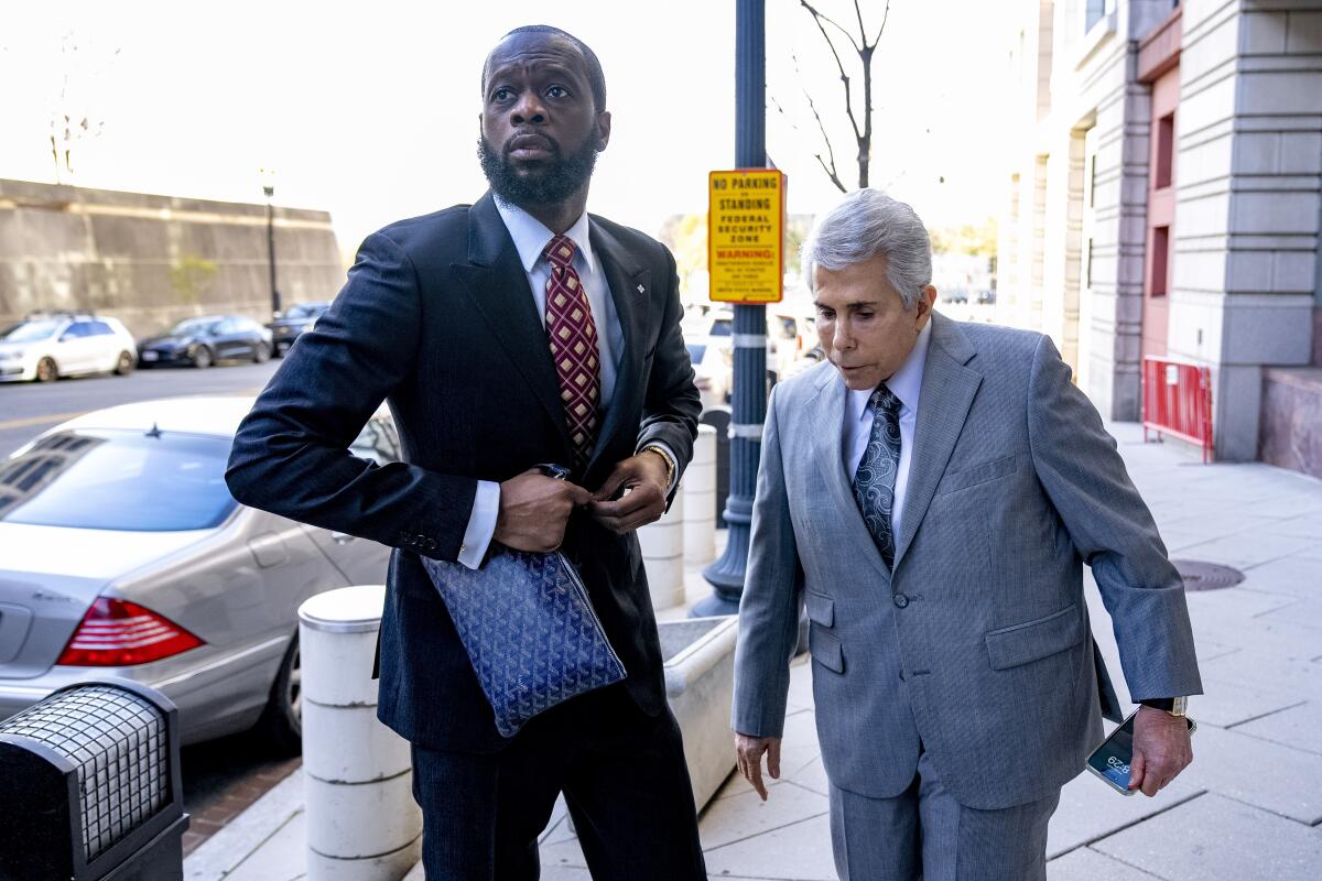 Pras buttons the jacket of his dark blue suit on a sidewalk as he looks away from lawyer David Kenner, who is in a gray suit