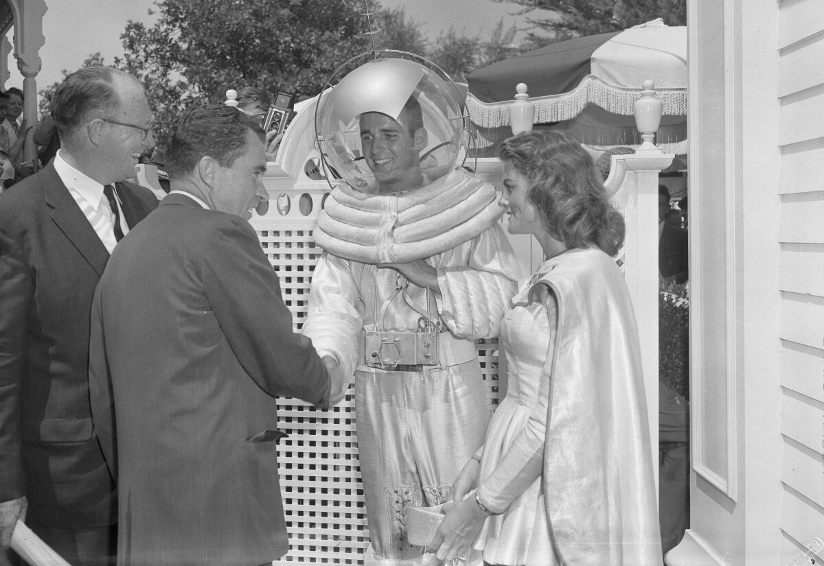 A man in white shirt and dark suit shakes the hand of a man in a clear, domed head cover standing by a woman in a cape.