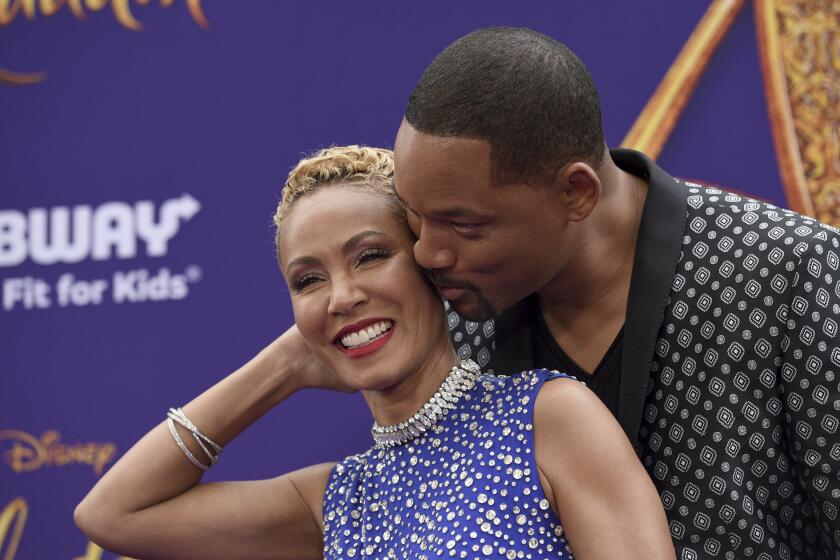 Jada Pinkett Smith in a blue, bejeweled top embraced by Will Smith in a black patterned suit jacket. He kisses her left cheek