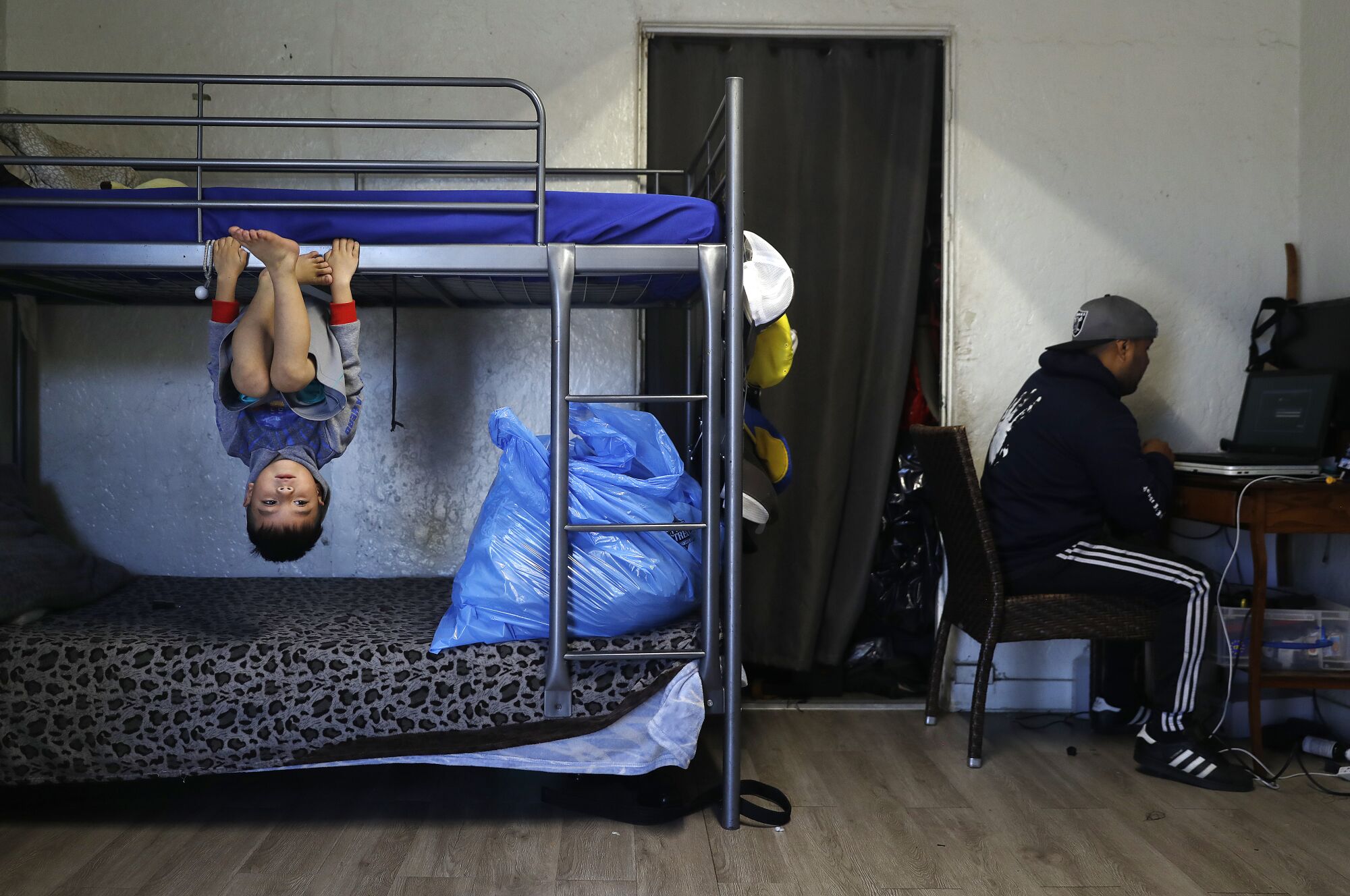 A boy hangs upside down on a bunk bed, while a man sits in front of the computer on the right