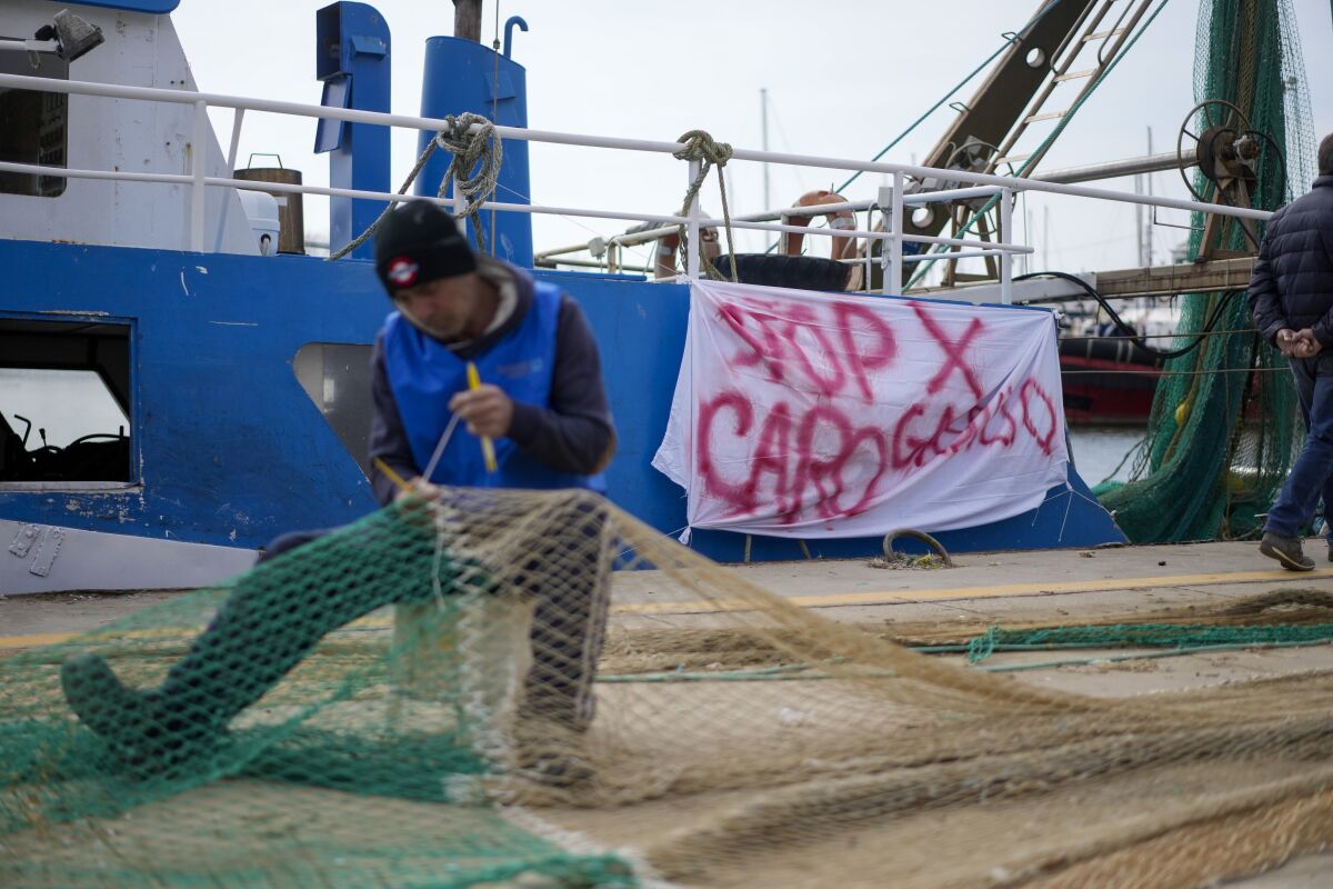 A fisherman mends a net in front of his fishing boat with a banner reading "stop for gasoline increase", in the Roman port of Fiumicino, Friday, March 11, 2022. Fishermen, facing huge spikes in oil prices, stayed in port, mending nets instead of casting them. Nowhere more than in Italy, the European Union’s third-largest economy, is dependence on Russian energy taking a higher toll on industry. (AP Photo/Andrew Medichini)