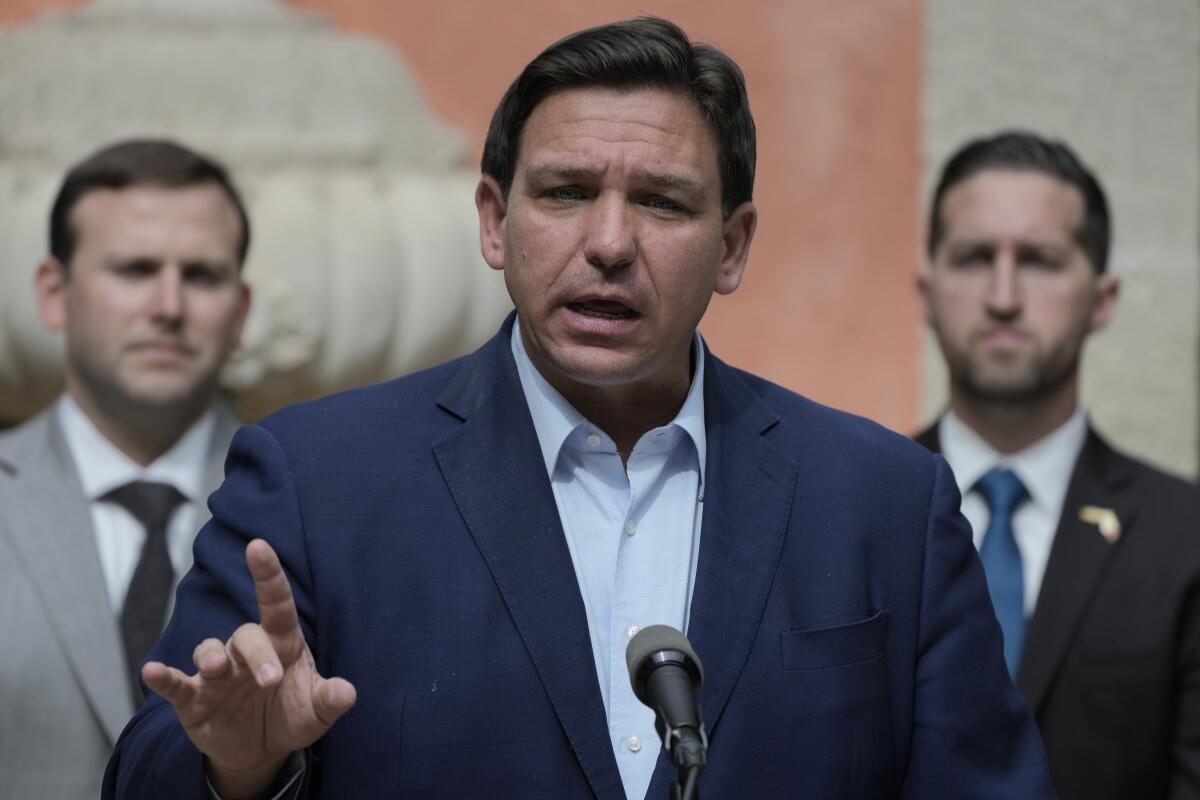 Ron DeSantis, wearing a light blue shirt and dark blue jacket, gestures as he speaks before a microphone 