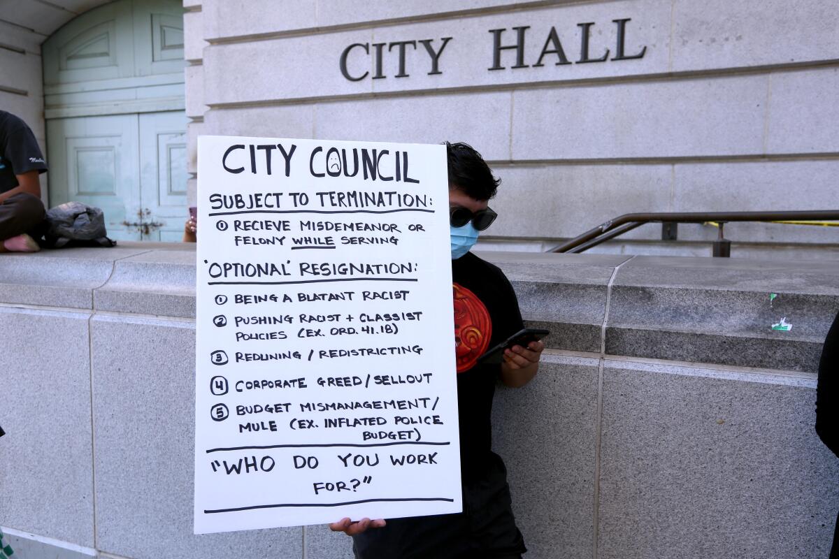 A demonstrator carries a sign listing "optional" reasons for councilmembers to resign, including "being a blatant racist."