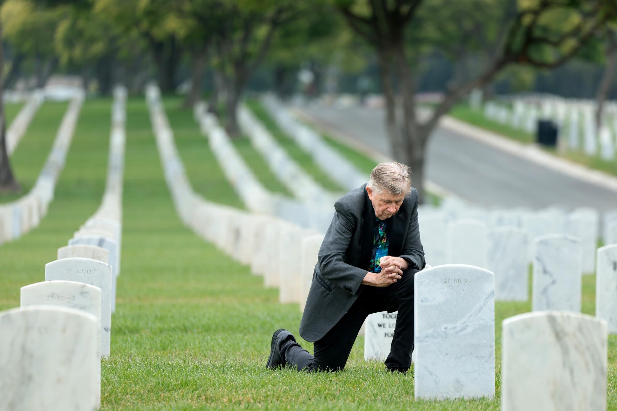 A man in a dark suit jacket kneeling in the grass, hands together, at one of hundreds of uniform white headstones