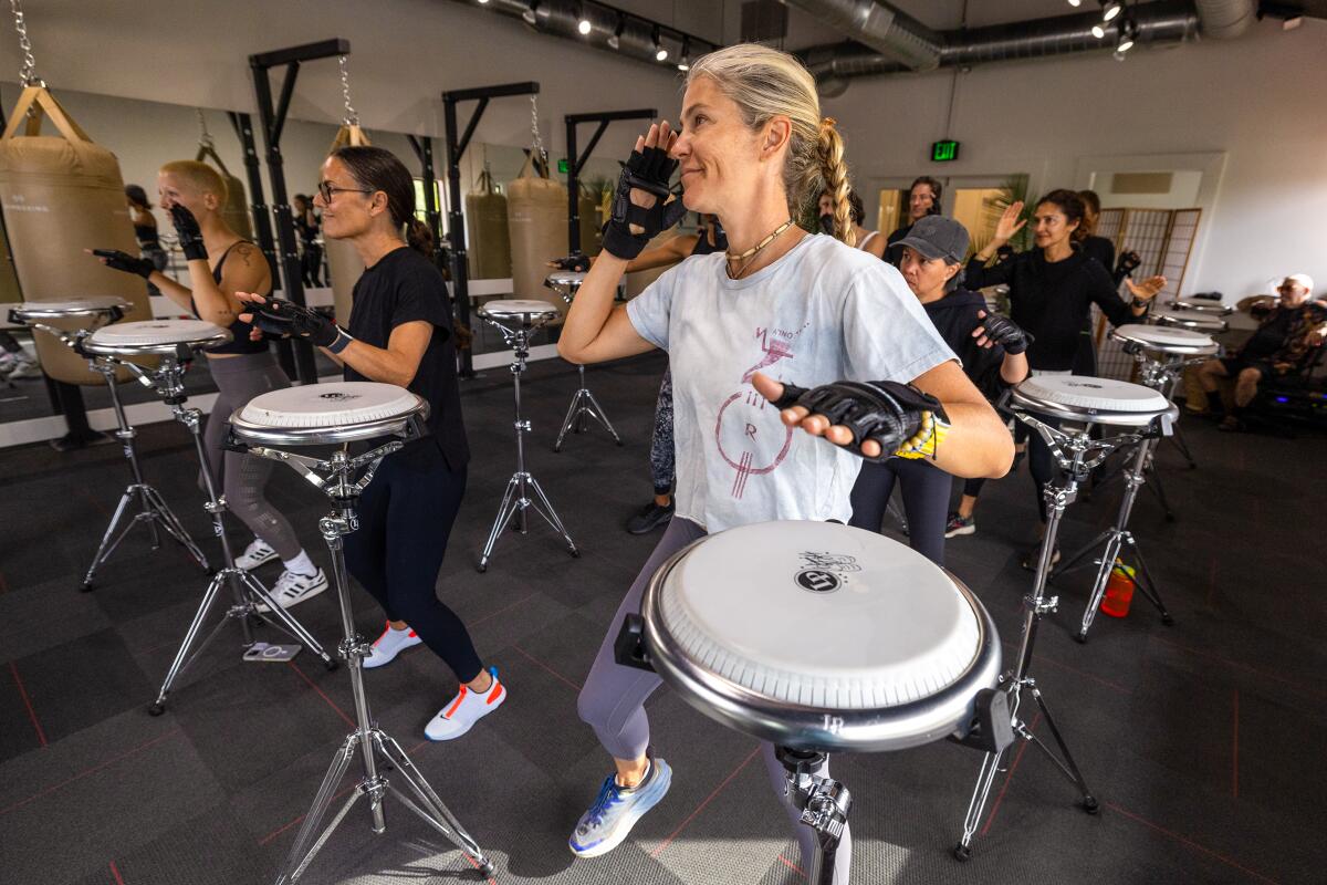 A woman wearing fingerless gloves touches her head while playing the drums during a drum boxing class. 