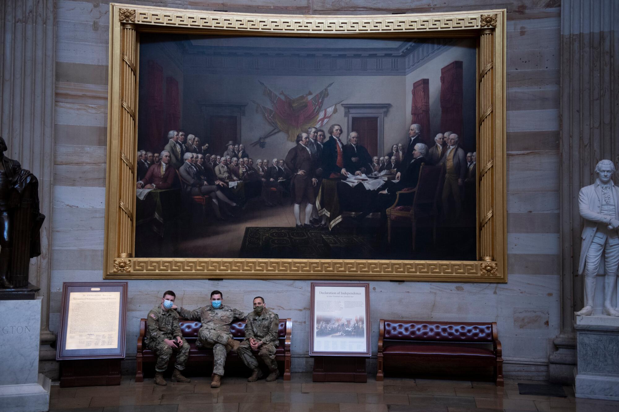 Three men in camo fatigues sit on a bench under a massive painting in the Capitol