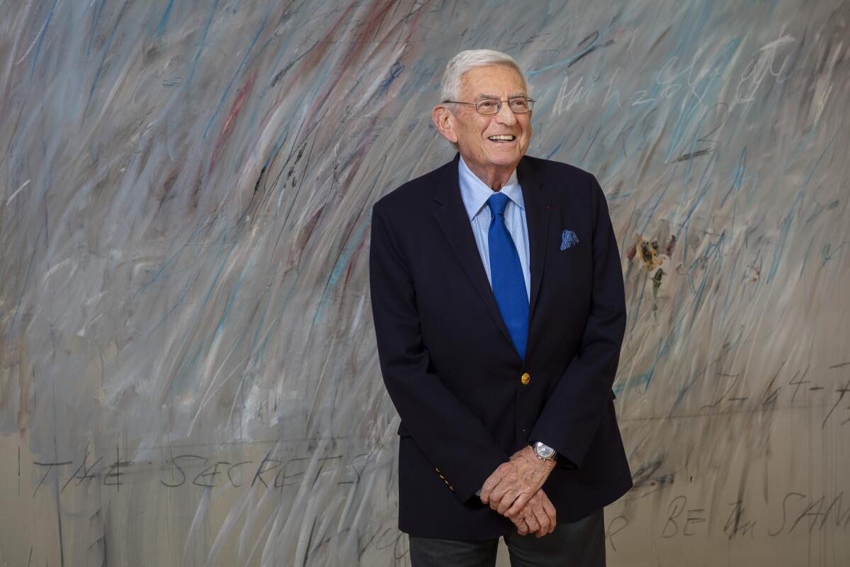 Eli Broad smiles and stands with his arms crossed in front of a large, gray-colored abstract painting 