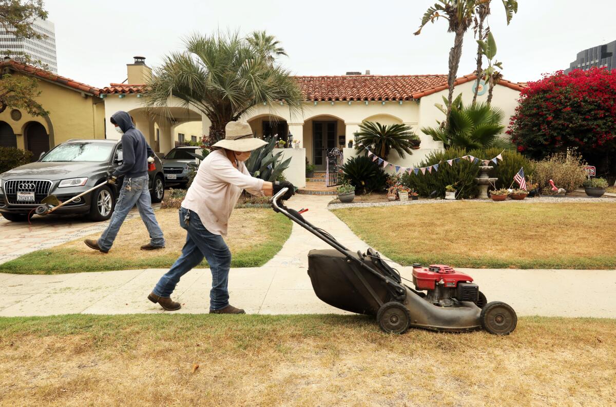 Workers use a weed trimmer and a lawnmower on a lawn that is mostly brown outside a Spanish-style home 