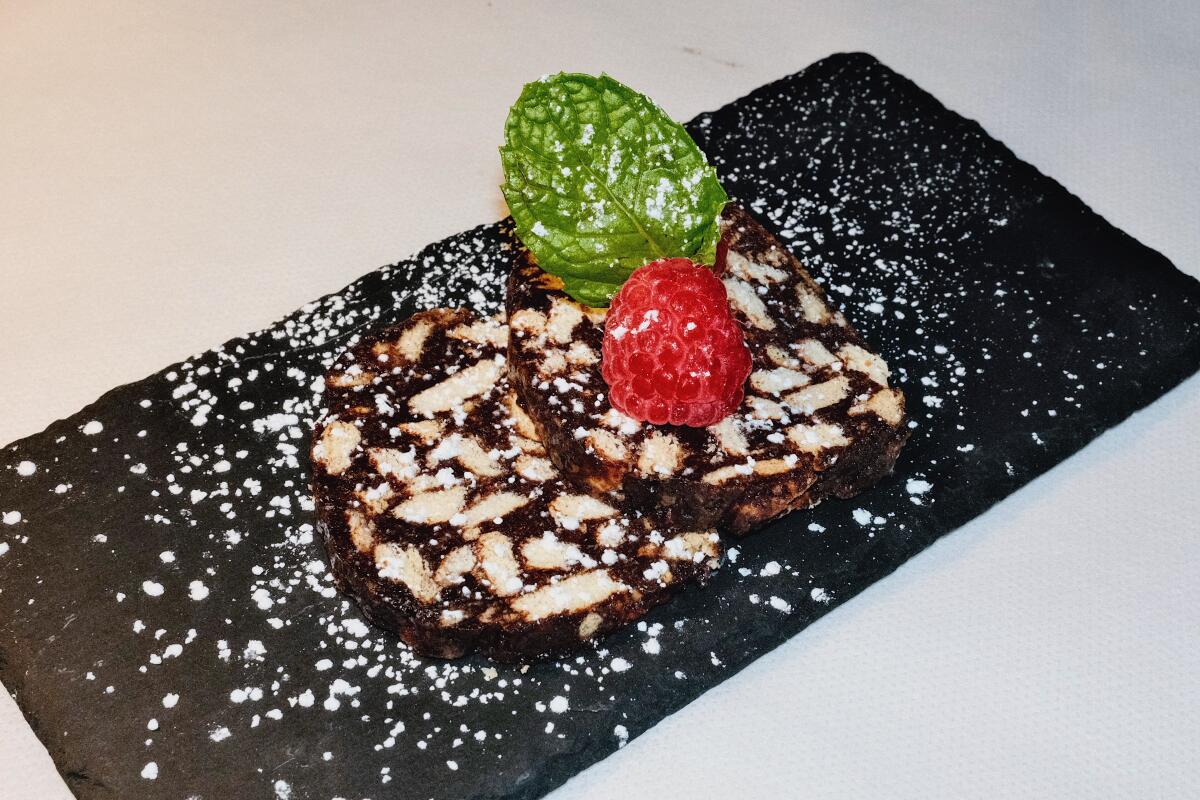 Two rounds of dark cocoa-powder cookies dotted with white cookie pieces on a black slate with powdered sugar and a raspberry