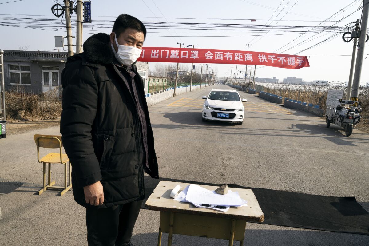 The street to a Chinese village is blocked as a man keeps track of people entering by using a thermometer and a paper list on the table.
