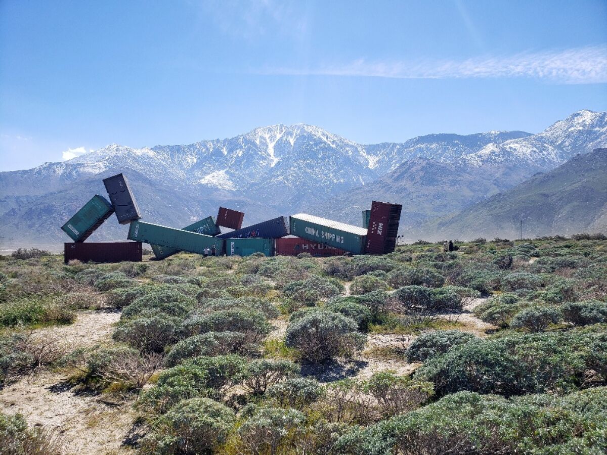 A sculpture that looks like wrecked freight train cars, with desert scrub in the foreground and snow-capped mountains behind