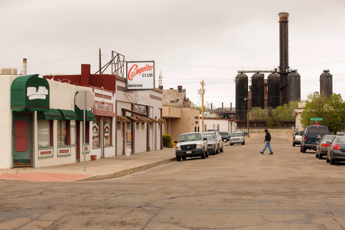 A person crossing a street lined with shops, passing in front of a steel mill at the end of the street.