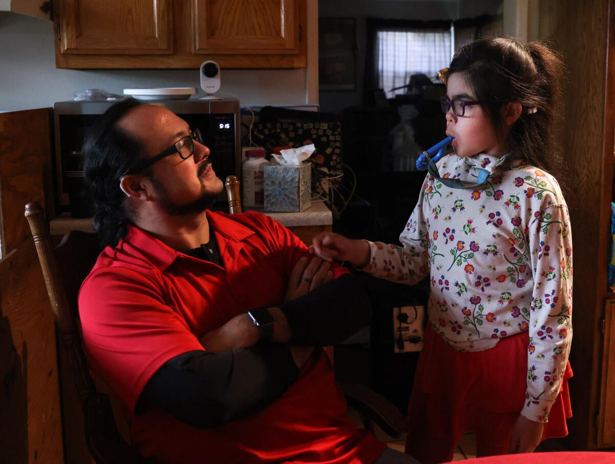 A man sits at a table while looking at his daughter, who holds a device in her mouth.