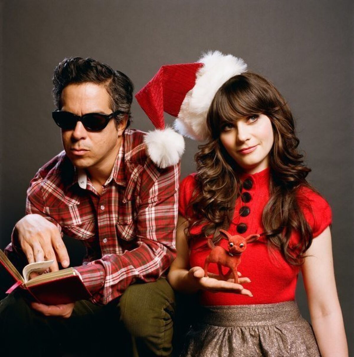 She & Him are M. Ward, in dark sunglasses and paging through a book, and Zooey Deschanel. wearing a Santa hat and holding a Rudolph figurine.