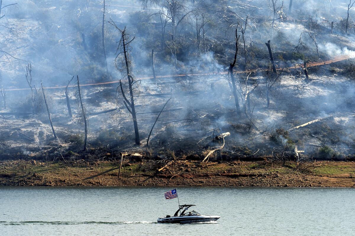 A boat crosses a lake as smoke rises from the charred hillside beyond the water