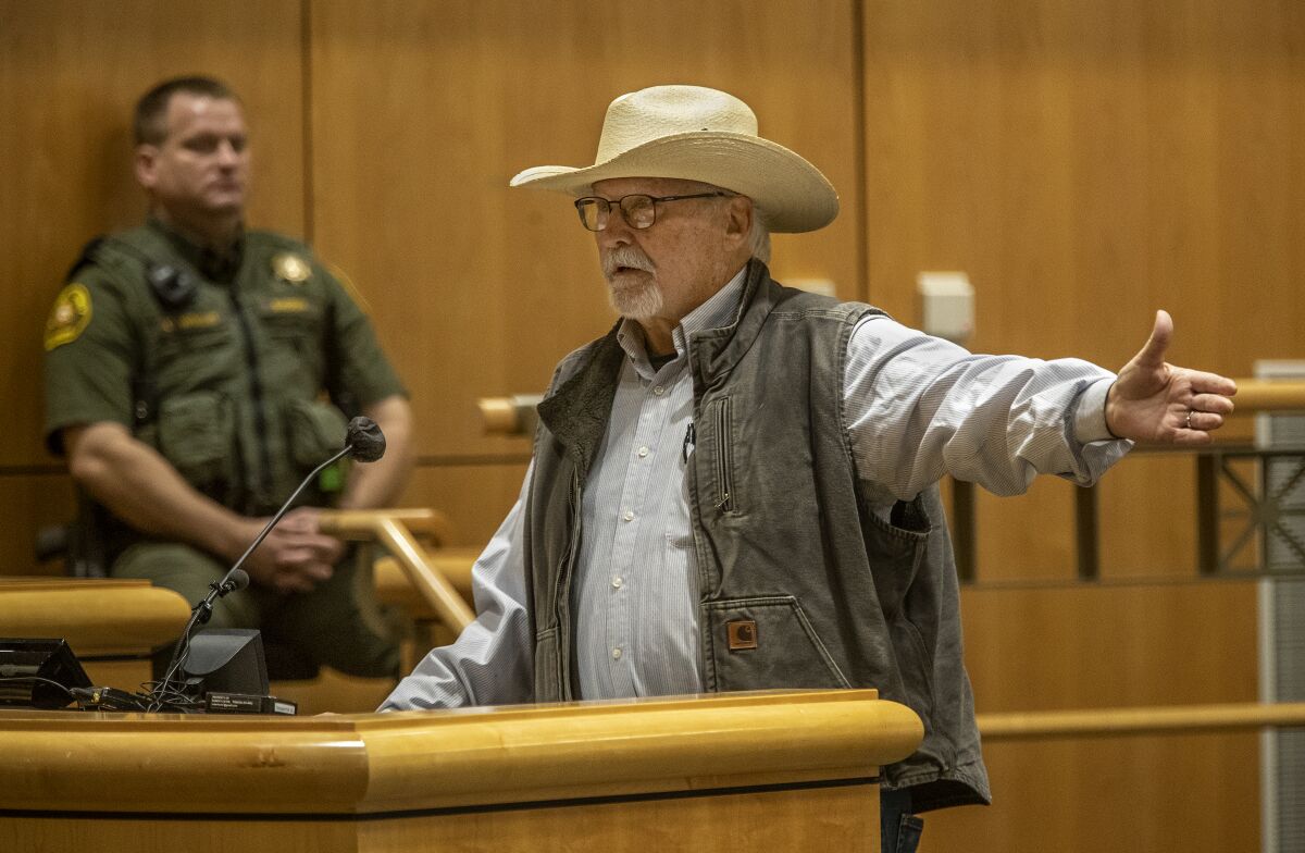 A Shasta County man speaking and gesturing before a lectern as a law enforcement officer watches.