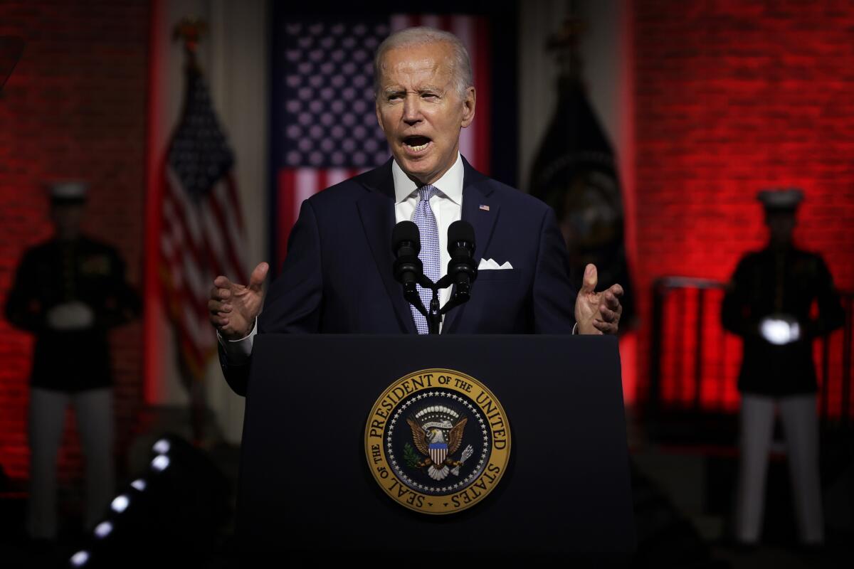 President Biden stands at a lectern with red lighting and the American flag in the background. 