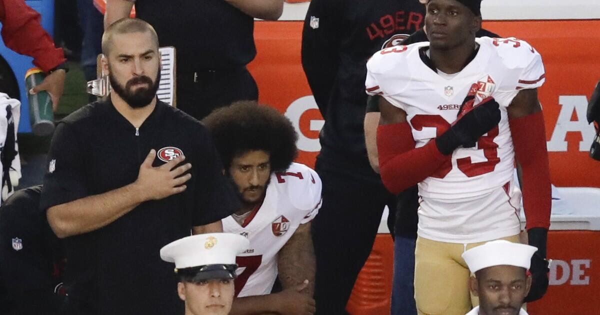 A man in a white football uniform is flanked by other people holding their hands over their hearts. 