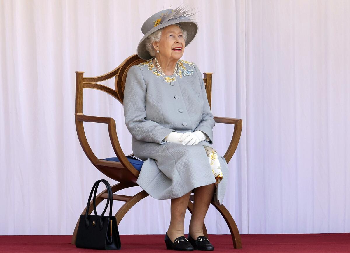 An elderly woman, seated, wearing a hat, looking up, gloved hands clasped on her lap, with a handbag by the chair.