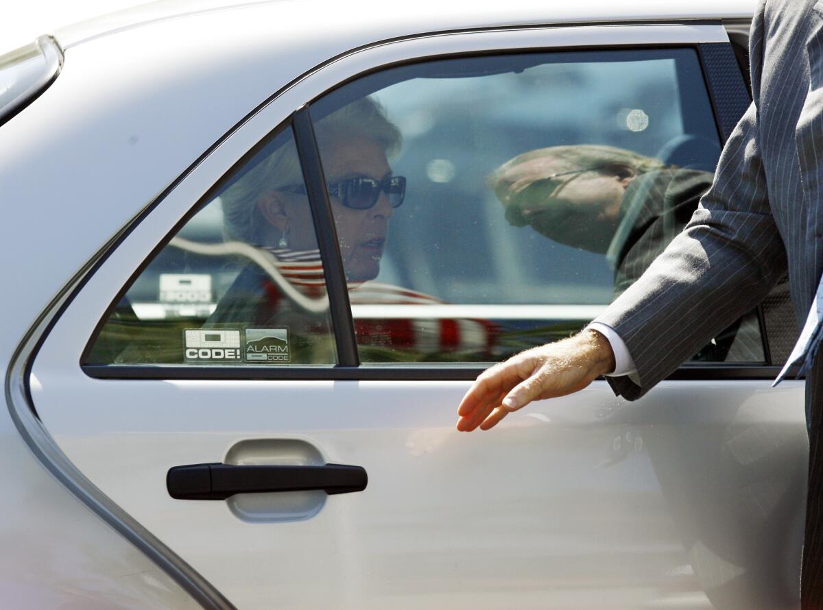A woman with short blond hair, sunglasses and dangly earrings sits in the back of a car.