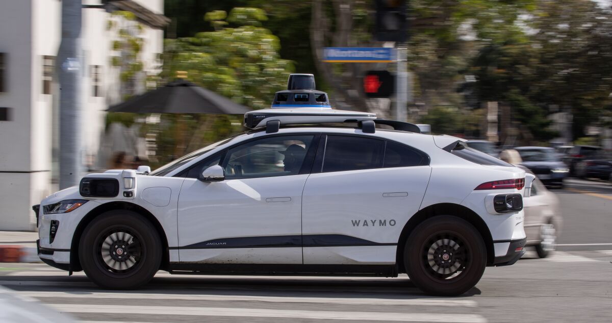  A white car with the word "Waymo" on its side and a self-driving sensor on its roof goes through an intersection. 